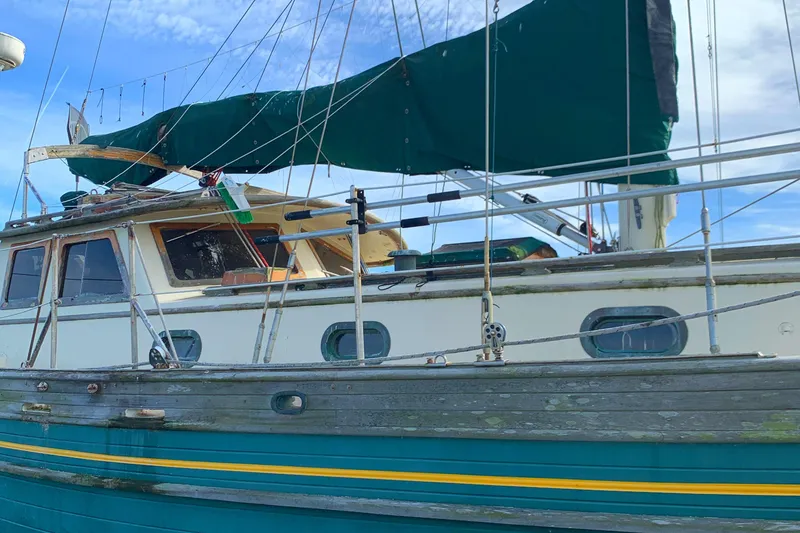 1980 Tayana Pilot House sailboat with green sails and wooden deck, docked under blue sky.