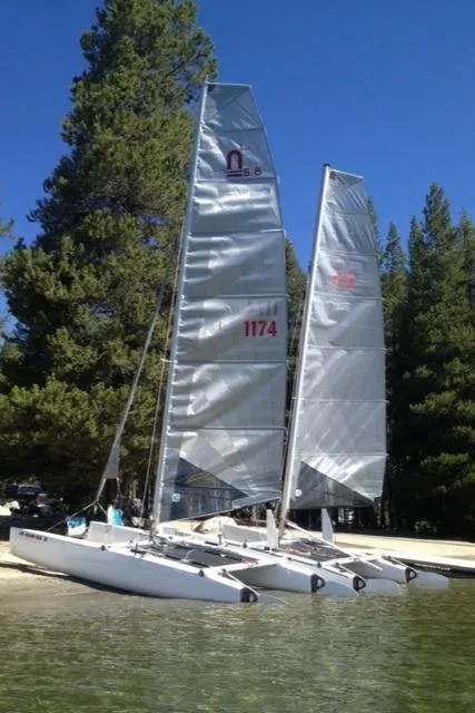 1989 Nacra 5.8 catamaran with sails, docked by a forested lakeside.