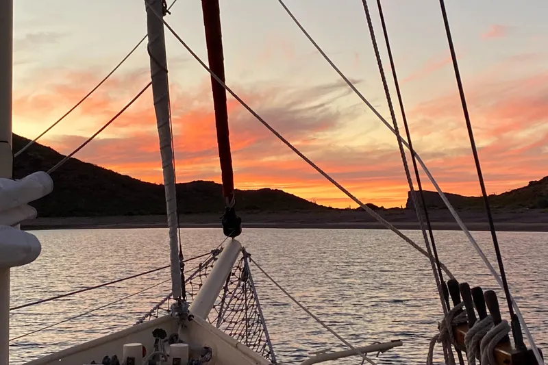 Schooner at sunset on calm waters, silhouetted hills, vibrant sky.