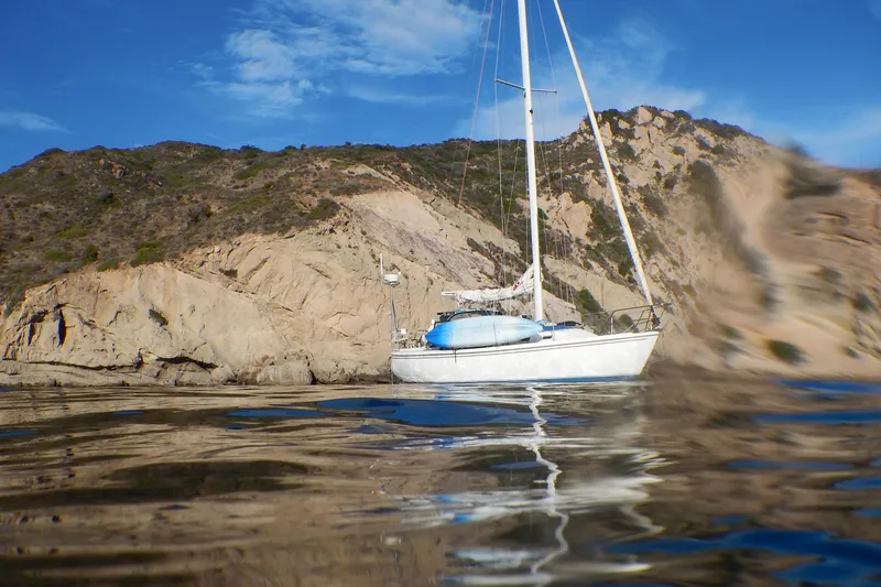 Catalina 34 sailboat anchored near rocky coastline, 1987 model, under clear blue sky.