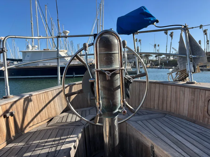 Steering wheel of a 1977 Baba 30 sailboat in a marina, with clear blue sky.