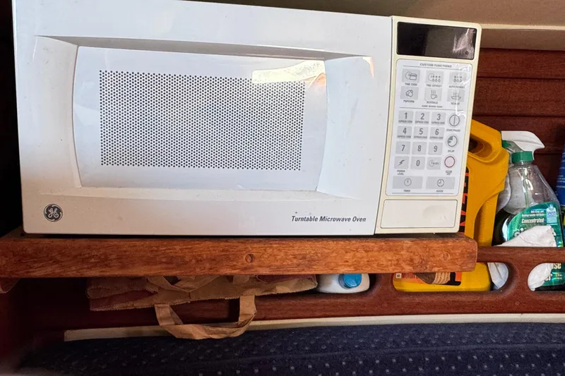 Microwave oven inside 1989 Ericson Sloop, surrounded by cleaning supplies and wooden shelving.